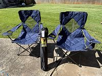 Two navy blue Berkey Jensen folding chairs with armrests and cup holders, outdoor on concrete with grass background