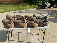 Eight duck decoys arranged on a folding table outdoors. Cork bodies, various painted wooden heads, two green mallard heads visible.
