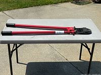 Photo shows a pair of 42-inch long bolt cutters with red handles and black grips, placed on a white folding table outside.