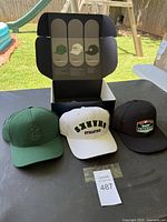 Set of three baseball hats (green, white, black) displayed in front of an open box with a tri-panel illustration inside.