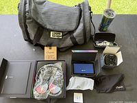 Gray fabric duffle bag, Origaudio headphones in packaging, blue PUFFER Bluetooth speaker in box, mini vacuum in black, insulated tumbler, and additional accessories