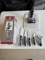 Photo showing one full set with 4 knives, peeler, and knife block on a table, items sealed in plastic except the block is unwrapped