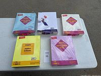 Five reams of Staples colored copy paper in various pastel and bright colors on a white table.