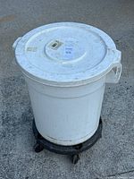 White commercial round trash can with lid on top and mounted on black dolly with caster wheels, showing use wear and dirt.