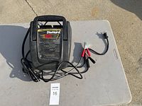 DieHard battery charger placed on a folding table outdoors showing the front panel with a digital display, indicator lights, connected clamps, and power cord.