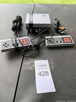 Photo showing the Family Computer System console with two wired controllers and the AV cable, all placed on a black outdoor surface.