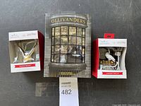 Three Harry Potter collectible items including the Golden Snitch ornament, Ollivanders display box, and Luggage Trolley ornament all new in box, arranged on a table with a MaxSold lot card number 482.