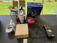 Overview of lot items on table including thermal tumblers, microwave rice cooker box, boxed Oster knife set, pink Crock-Pot, wire fruit basket, and taco holder.