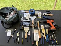 Overview of lot laid out on table showing various hand tools, scrapers, hammers, pliers, paintbrushes, power drill, tape, and electrical covers.