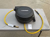 Black Husky retractable air hose reel placed on a gray folding table outside, yellow hose extending out with metal connectors on hose ends. Mounted design visible on side with mounting points.