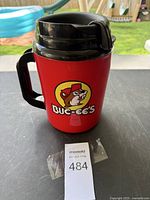 Front view of large red travel mug with black lid and Buc-ee's beaver logo on the front, sitting on a table outdoors.