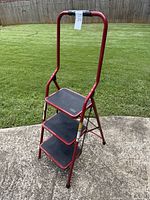 Front side view of red metal step ladder standing open on concrete surface with grassy background, showing three black textured steps, red frame and safety handlebar.