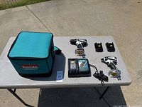 Full contents of the lot displayed on table outdoors: teal and black Makita soft case, cordless drill, impact driver, charger, two batteries (one inside tool, one alongside), and auction lot tag.