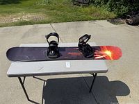 Morrow Torch snowboard with black bindings facing up on white folding table outdoors on concrete surface.