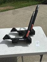 Photo showing two Yakima kayak racks placed on a white table outside on pavement. Both racks have black bases with Yakima branding and arms padded in black with red accents.