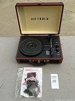 Full view of Victrola portable turntable in brown case with lid open. Case has a small nick on the exterior. Manual and power adapter in plastic bag shown.