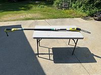 Full length side view of the Ryobi electric pruning saw with extended pole set on a table outside.