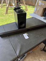 Samsung soundbar and subwoofer placed on a table outdoors, showing full length of soundbar and subwoofer front with cables on top.