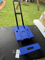 Blue small collapsing dolly fully extended on black table outdoors with grass background.