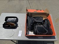 Snowboard boots and goggles set displayed on a table outside with boots inside an orange box, goggles inside a black hard case next to the box