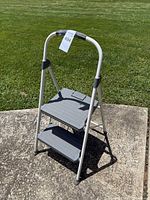 Two-step aluminum step ladder in open position on concrete with grass background, showing size and condition.