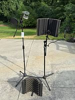 Wide view of two microphone stands outdoors on concrete surface with green background. Each stand features curved black foam acoustic shields. One stand has a pop filter and spider shock mount holding an MXL microphone.