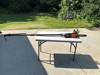 Full length view of Worx electric pruning saw with telescopic pole on folding table outdoors, showing entire tool including handle and extension pole.
