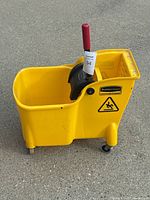 Yellow Rubbermaid mop bucket with black wringer and red handle, showing side caution slip hazard sticker, on caster wheels.