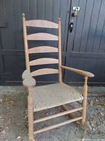 Full view of tall antique wooden rocking chair with woven seat and visible wear, including missing parts on the seat and one repaired back slat.