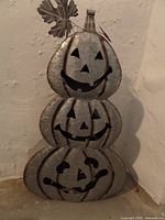 Front view of a three-tiered stacked metal pumpkin decoration showing carved jack-o'-lantern faces and a decorative metal leaf on top.
