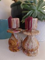 Front angled view of two onyx candle holders featuring swirls of brown, beige, and red colors, placed in front of plants and a blue pot.