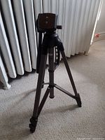 Camera tripod standing with legs extended on carpet floor next to heater