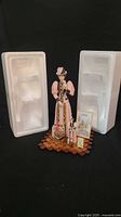 Full view of the larger figurine standing between two clear Styrofoam packaging pieces showing all included items: large and small figurines, two advertising signs, two small bags, and wooden base.