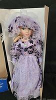 Porcelain doll wearing lavender and purple Victorian-style dress with lace accents, standing inside original box.