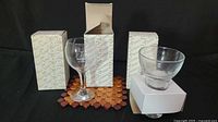Set of three clear glass wine glasses and one glass candle stand displayed with their original boxes on a black background.