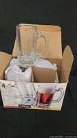 Set of four clear glass beer stein mugs with large handles, inside and on top of the original printed box. One mug is placed outside the box for display.