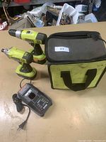 Two Ryobi cordless drills, a battery charger and a soft carry bag placed on a table, showing the entire kit.