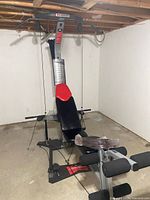 Bowflex home gym machine including bench with red and black cushioning, resistance rods, and pulley system, situated in a basement with unfinished ceiling and concrete floor.