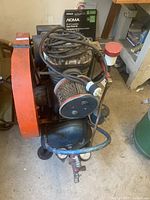 Angle view of the Leland air compressor with visible orange cooling fan cover, compressor head with hoses coiled on top, and black motor housing on a cart system.