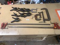 Photo showing all clamps laid out on a table: 3 metal C clamps, 7 black plastic spring clamps, and 2 large red metal bar clamps.