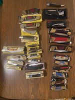 Overview of 28 manual fold‐out pocket knives on wood floor, includes Frost Cutlery, Bear Hunter and other brands