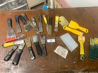 Top-down photo showing 30 scrapers arranged on a brown table, 20 metal with various blade widths and handles, and 10 plastic yellow and white scrapers.