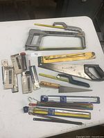 Wide photo showing two hacksaws, one hand saw, and multiple packs of replacement blades arranged on a white surface.