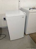 Full view of the white Sanyo mini fridge positioned beside another appliance on a grey floor, showing the closed door and Sanyo logo.