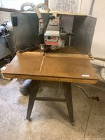 Full view of the vintage DeWALT radial arm saw mounted on a wooden table with metal legs and dust collector hose attached.