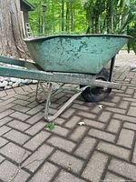 Side view of green metal wheelbarrow showing rust and paint wear, flat front tire visible.