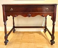 Front view of dark wood antique desk showing drawer, scalloped apron, turned legs, casters