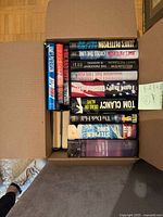 Top-down view of open cardboard box containing 28 novels showing visible spines of Patterson, Clancy, King, etc.
