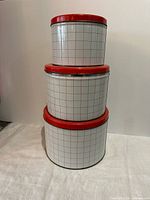 stacked view of three white canisters with red grid and lids
