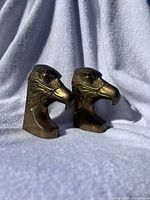 Two brass eagle head bookends standing side by side on a white textured fabric background.